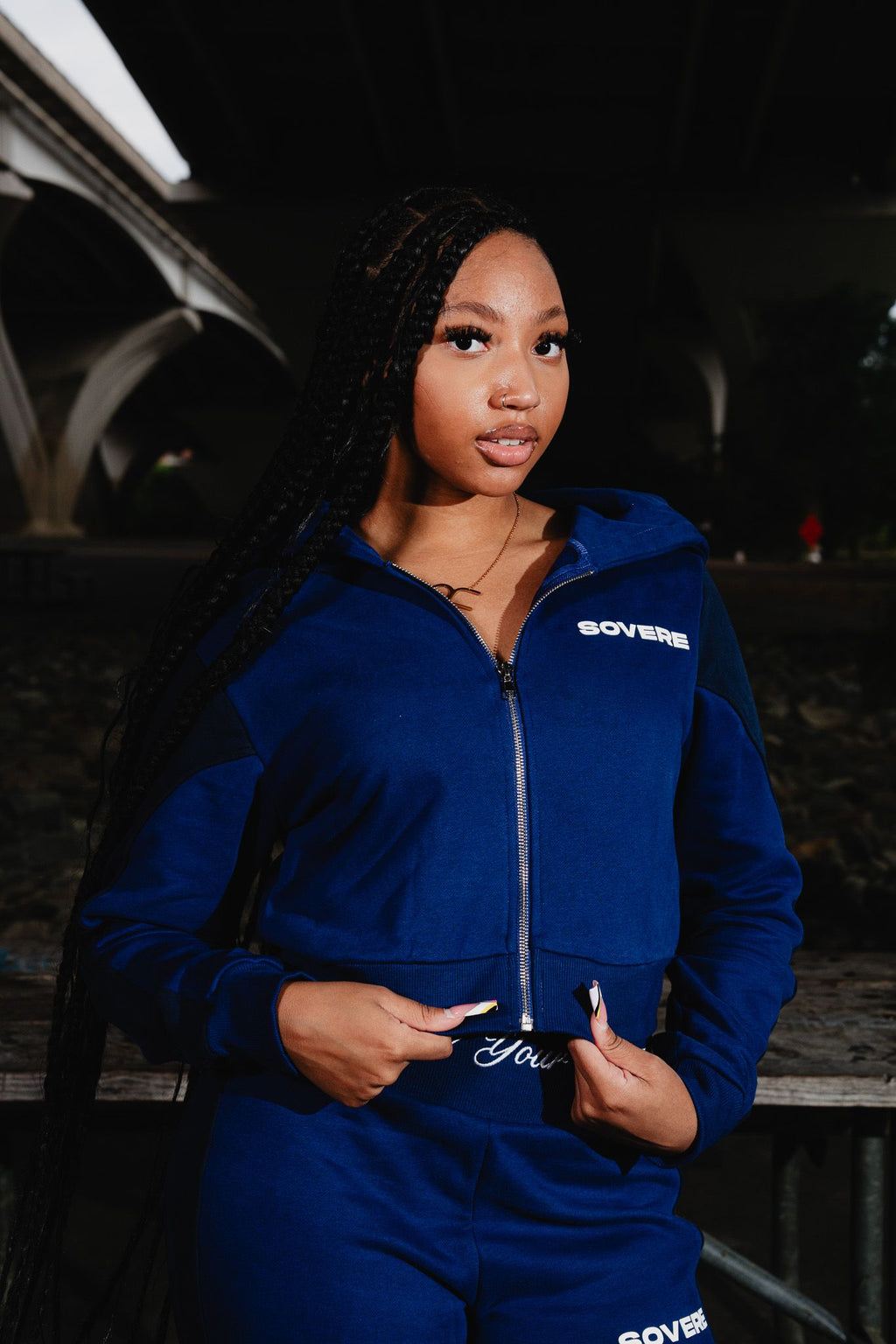 A woman with long black braids wearing a Medium navy-blue zip-up hoodie and matching sweatpants with 'SOVERE' printed in white on the chest, photographed outdoors at night under a bridge.