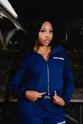 A woman with long black braids wearing a Medium navy-blue zip-up hoodie and matching sweatpants with 'SOVERE' printed in white on the chest, photographed outdoors at night under a bridge.