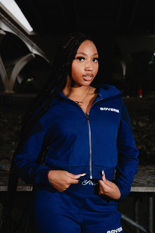 A woman with long black braids wearing a Medium navy-blue zip-up hoodie and matching sweatpants with 'SOVERE' printed in white on the chest, photographed outdoors at night under a bridge.